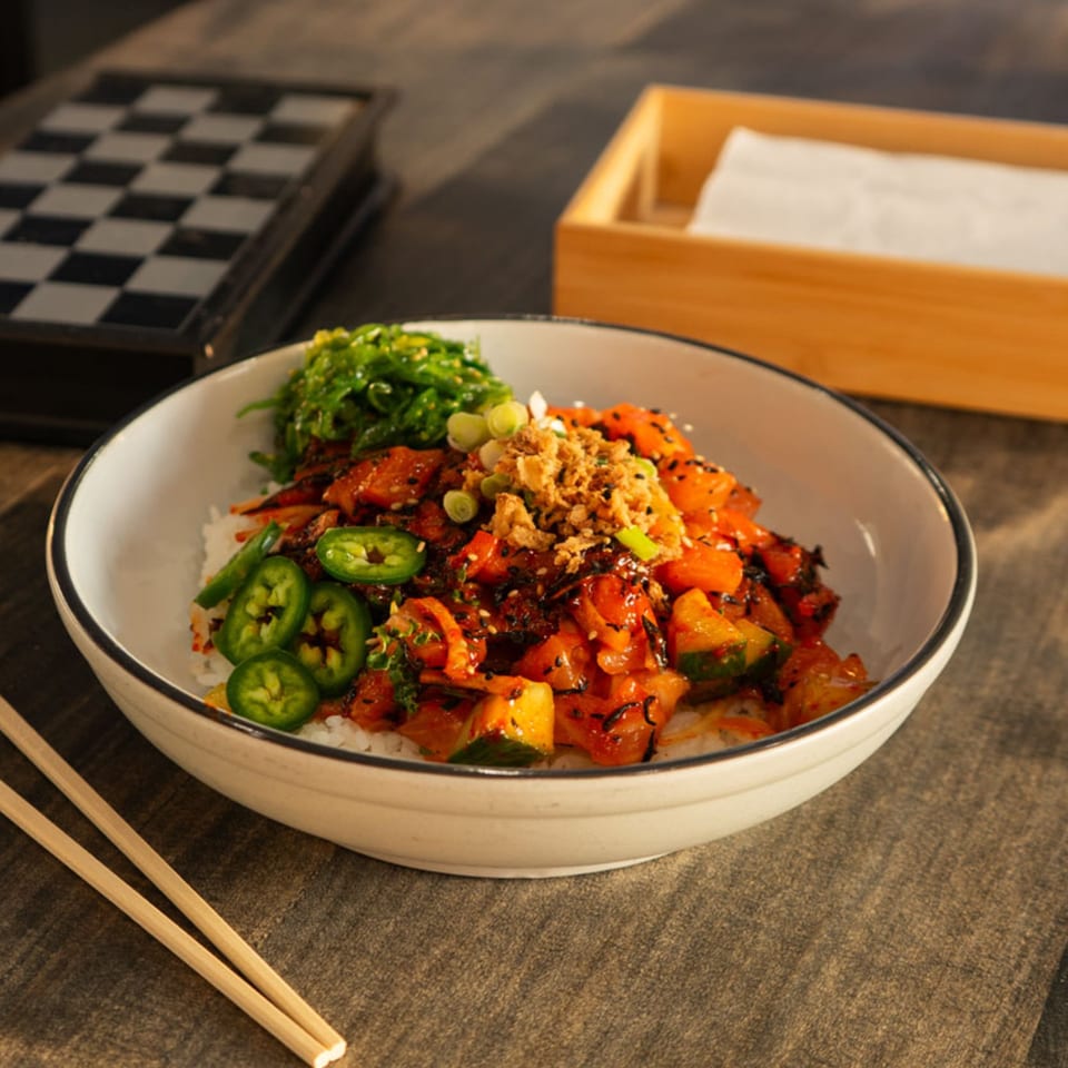 A colorful Hana Poke bowl with salmon, seaweed, jalapeños, and crispy onions, with chopsticks nearby.